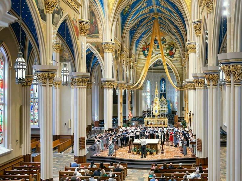 A high-angle view of the Basilica of the Sacred Heart at the University of Notre Dame. A choir stands on a stage at the front of the church, facing the altar. The conductor stands with his back to the audience, directing the choir. The audience sits in wooden pews, facing the stage. Sunlight streams in through stained-glass windows along the sides of the church. Tall white columns with gold accents line the nave. The vaulted ceiling features intricate blue and gold details, as well as religious murals.