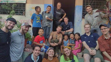 A group of Notre Dame students smiles and poses with local children on the steps of a brightly painted building in a Latin American community. Some children hold small paper cups.