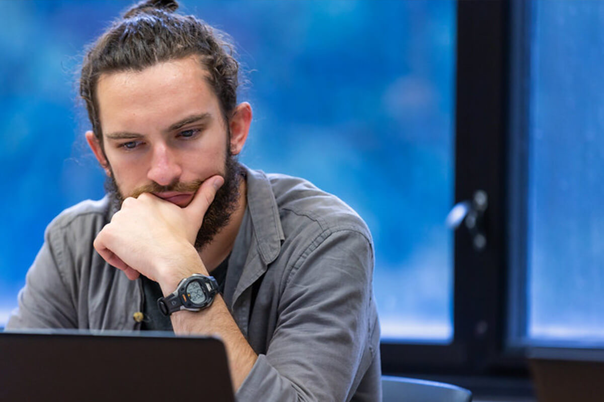 A student with a man-bun and a beard rests his chin on his hand as he looks thoughtfully at his laptop.