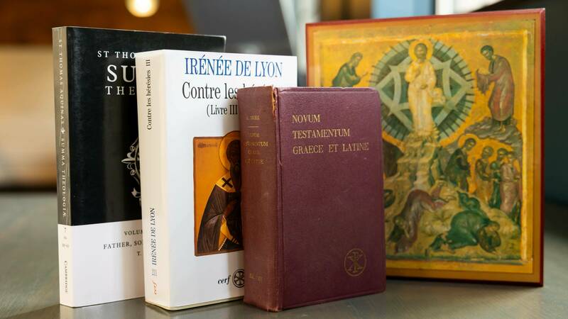 Four theological books and an icon rest on a gray surface.  From left to right: a black Summa Theologica, a white book titled "IRÉNÉE DE LYON Contre les hérésies III", a burgundy "Novum Testamentum Graece et Latine," and a framed icon depicting a gold-robed figure surrounded by other people.